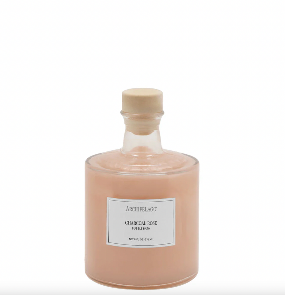 Product photograph by Archipelago featuring an apothecary glass bottle of Charcoal Rose Bubble Bath with a minimalist brand label and light wood stopper filled with peach liquid on a white backdrop