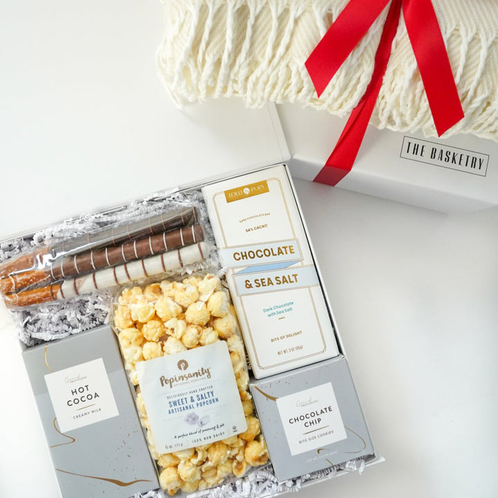 Product photograph by The Basketry of a white gourmet gift box featuring Popinsanity popcorn, chocolate-dipped pretzels, and a sea salt chocolate bar next to a cream knit blanket tied with a vibrant red ribbon