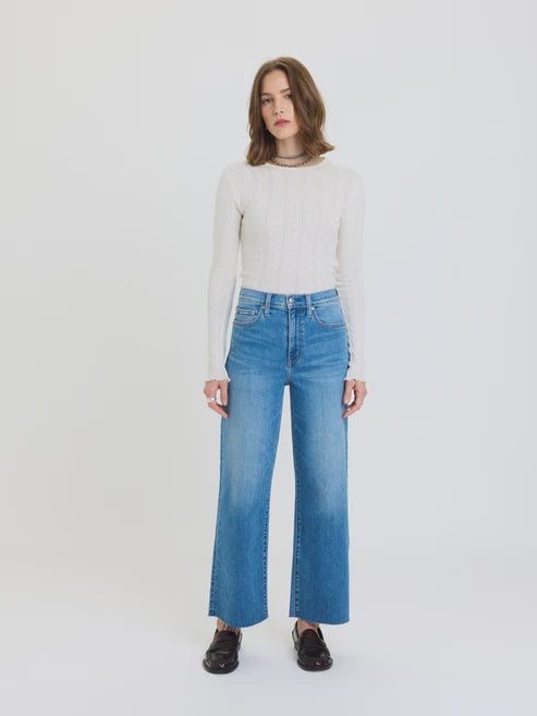 Studio photograph of a woman in a bright minimalist studio wearing a white pointelle long-sleeve top, high-waisted wide-leg blue denim jeans with raw-edge hems, and dark leather loafers