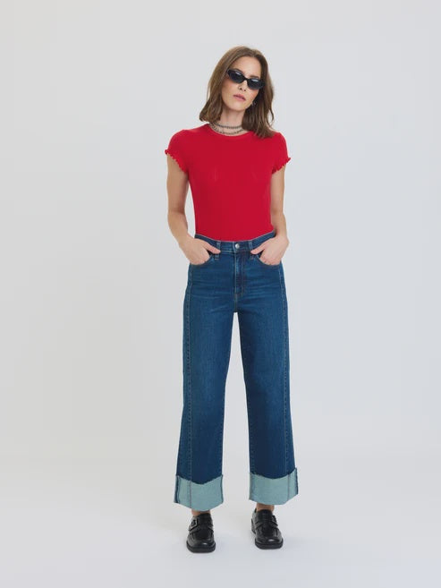 Studio fashion photography of a woman wearing a vibrant red lettuce-edge top, dark blue wide-leg jeans with oversized light-blue cuffs, and black buckled loafers against a plain white backdrop
