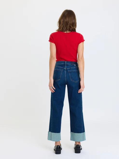 Studio photograph of a woman from behind against a white background wearing a vibrant red lettuce-edge t-shirt and dark blue wide-leg jeans with distinctive light blue cuffed hems and black loafers
