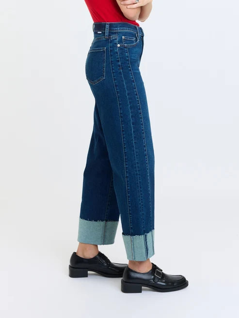 Commercial studio photograph of a model wearing high-waisted dark denim jeans featuring oversized light-wash raw-edge cuffs and polished black leather buckle loafers against a minimalist white backdrop