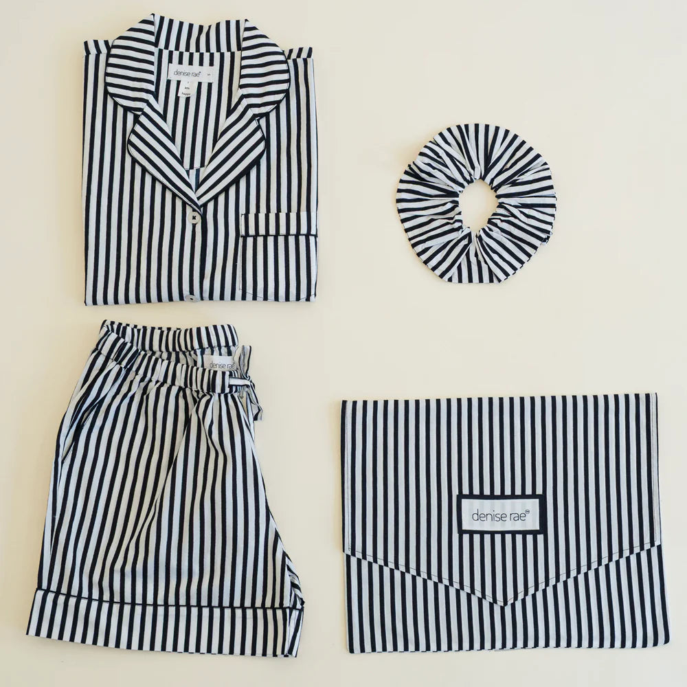 Flat-lay product photography by Denise Rae featuring a black and white vertical striped loungewear set including a notched-collar shirt, matching shorts, a scrunchie, and an envelope pouch on a minimalist neutral background