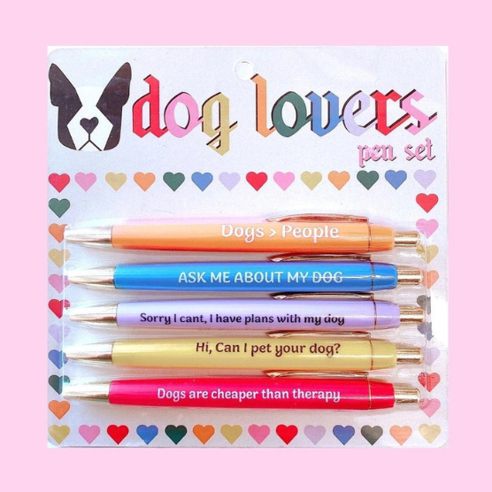Stationery product photography of a dog lovers pen set showcasing five colorful pens in orange, blue, lavender, yellow, and pink featuring humorous dog slogans and a French bulldog icon on heart-patterned packaging board against a soft pink background