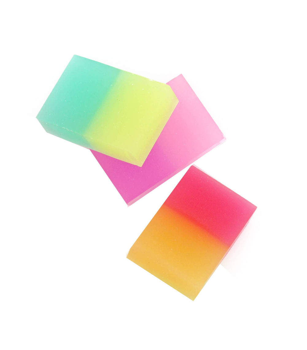 Product photograph of three rectangular, translucent glycerin soap bars arranged on a white background featuring vibrant color gradients of green to lime yellow and bright orange to red-pink alongside a solid magenta bar