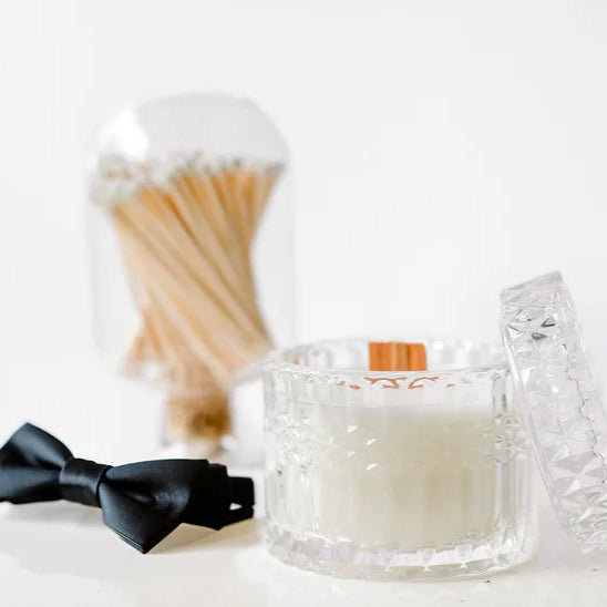 Photograph of a white candle with a wooden wick in a faceted glass jar positioned next to a black satin bow tie and a glass cloche filled with white-tipped matches on a minimalist white surface