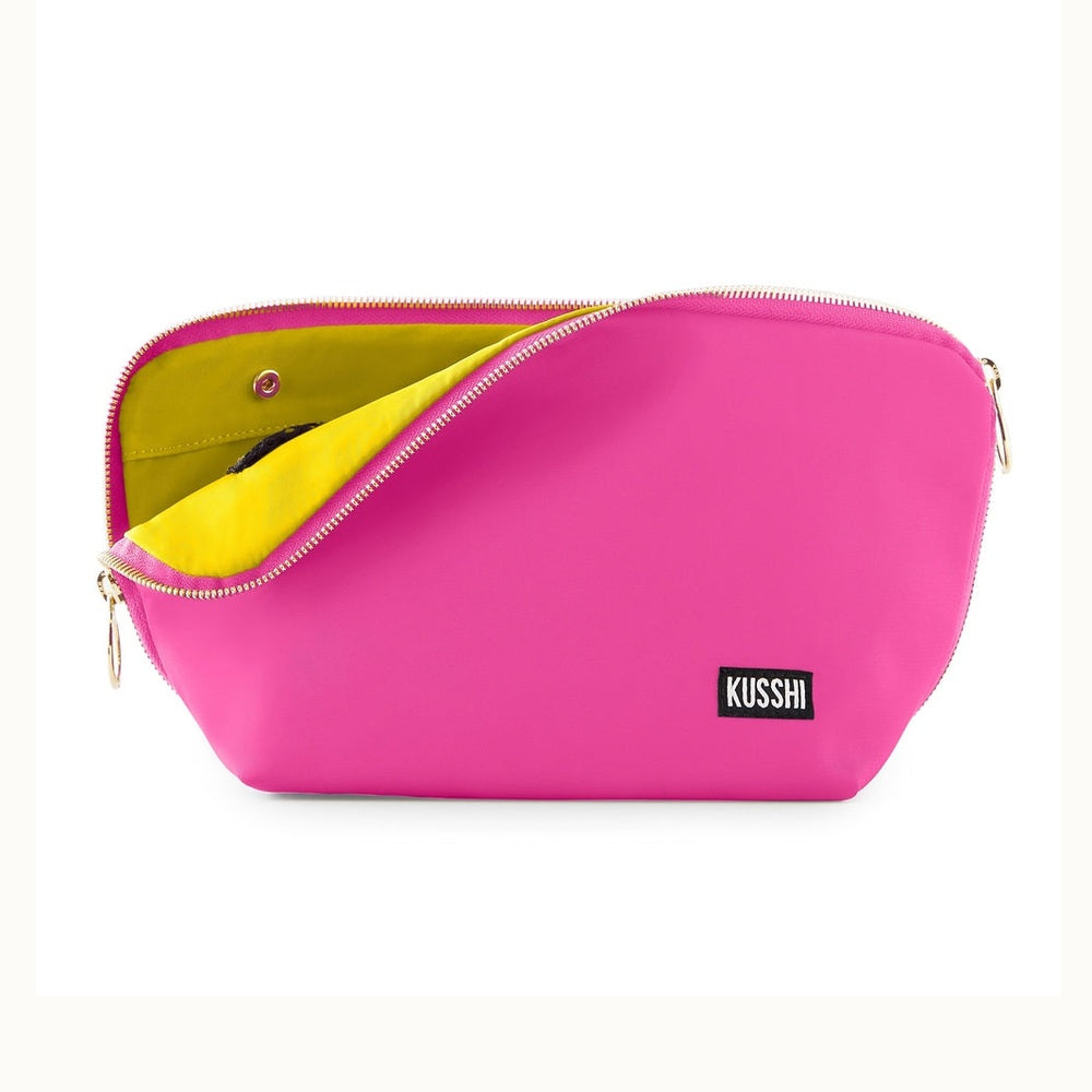 Product photograph by Kusshi of a hot pink makeup bag in a studio setting featuring a contrasting lime green lining, gold-toned zipper pulls, and a black brand label