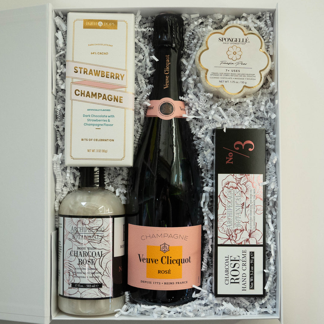 Photograph of a luxury gift box featuring a central bottle of Veuve Clicquot Rosé Champagne with a salmon-pink label nestled in white shredded paper alongside Archipelago Botanicals skincare and a flower-shaped Spongellé buffer