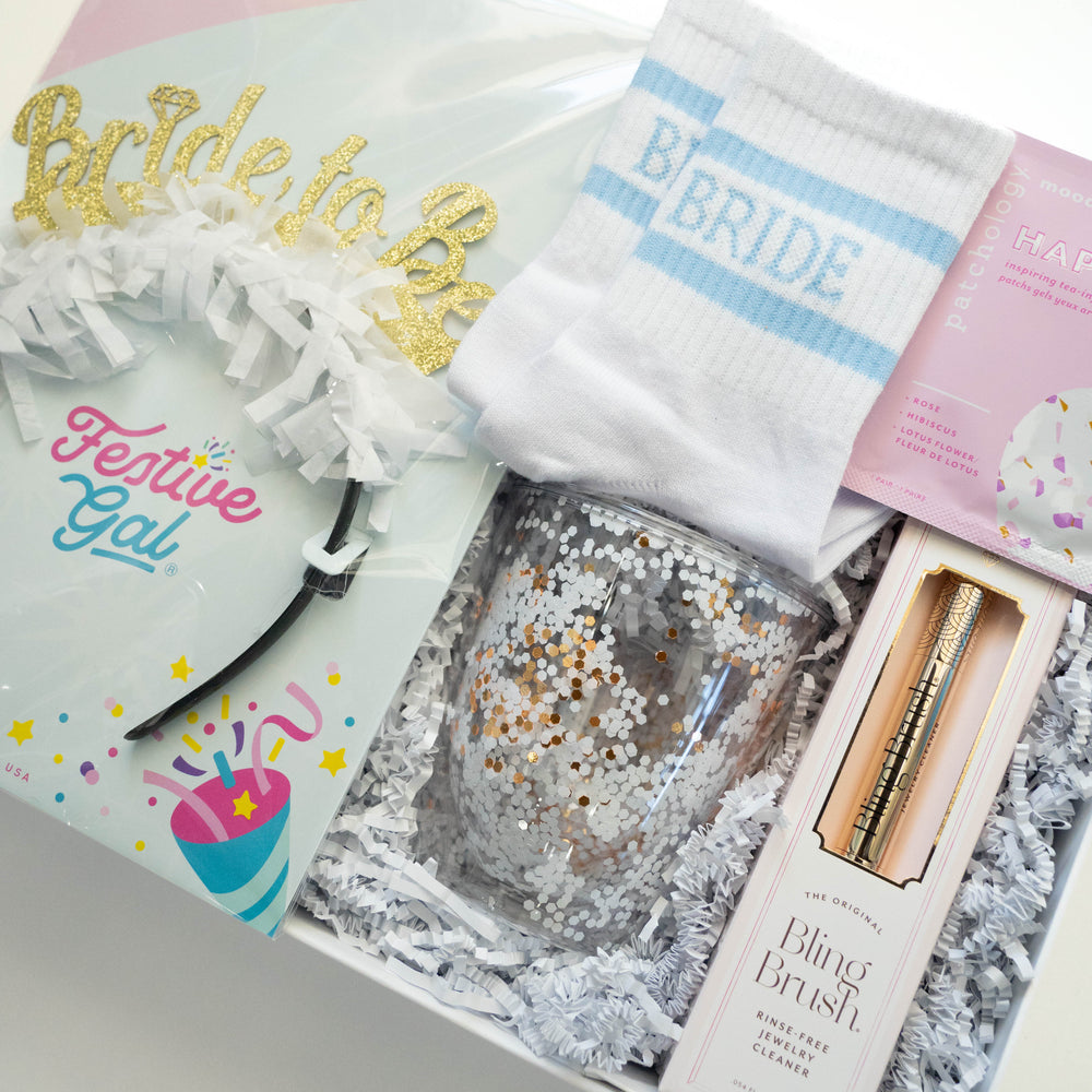 Photograph by Festive Gal of a bridal gift box featuring a gold glitter Bride to be headband, blue striped socks, a shimmering white and gold confetti tumbler, and a Bling Brush jewelry cleaner nestled in white shredded paper