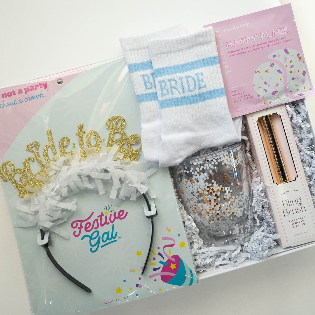 Photograph of a Festive Gal bridal gift box featuring a gold glitter Bride to Be headband, white socks with light blue stripes, and a pink Patchology eye gel packet on white shredded paper