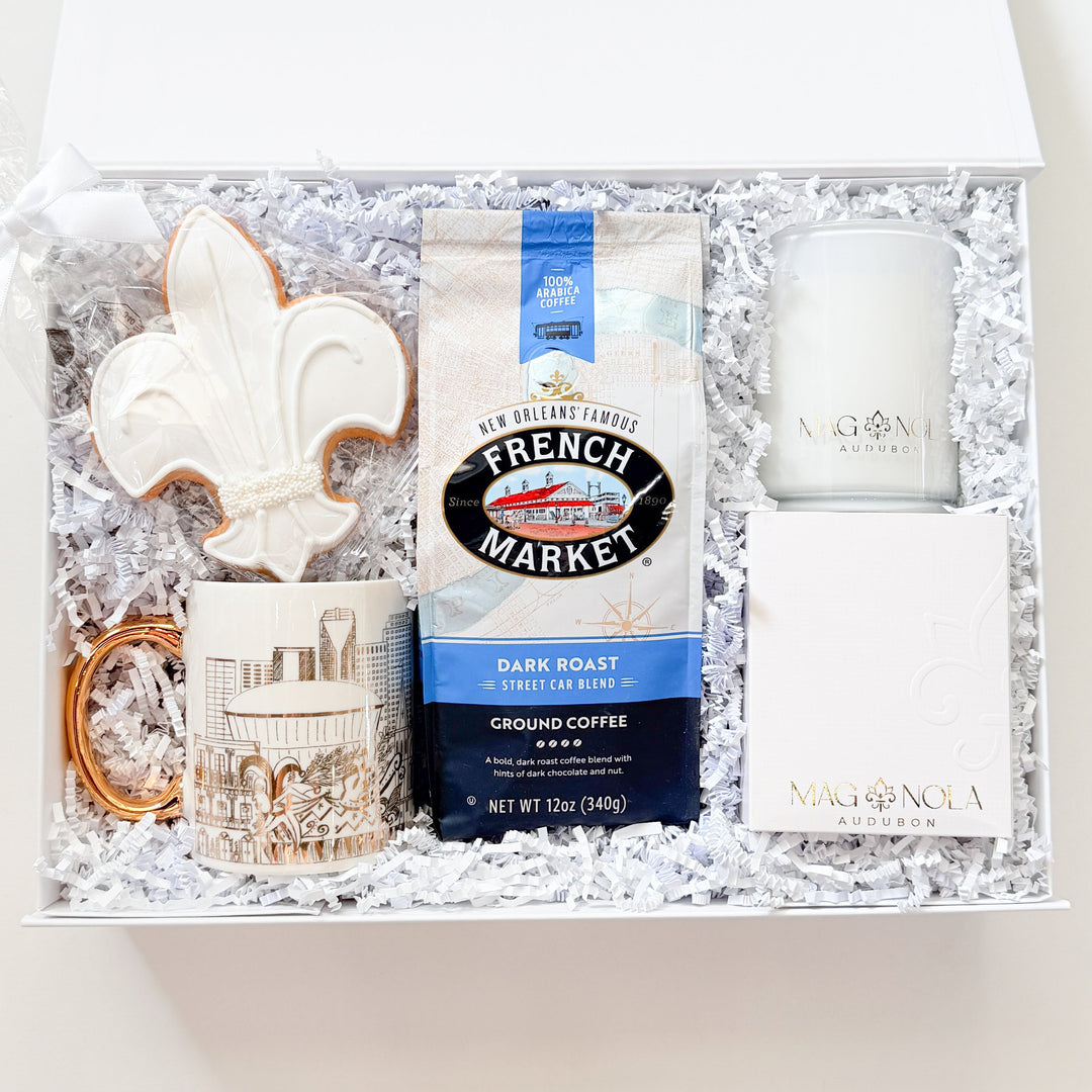 Photograph of a Magnolia Audubon New Orleans themed gift box featuring a large white iced fleur-de-lis cookie with pearl accents, a bag of French Market coffee, and a gold-handled mug showcasing the Caesars Superdome nestled in white crinkle paper