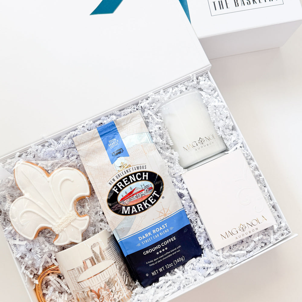 A curated gift box set from The Basketroy, featuring a white gift box with a teal ribbon, an open box filled with white crinkle paper and assorted gift items including a French Market coffee bag, a white candle, a white soap bar, a ceramic mug, and a white decorative piece.