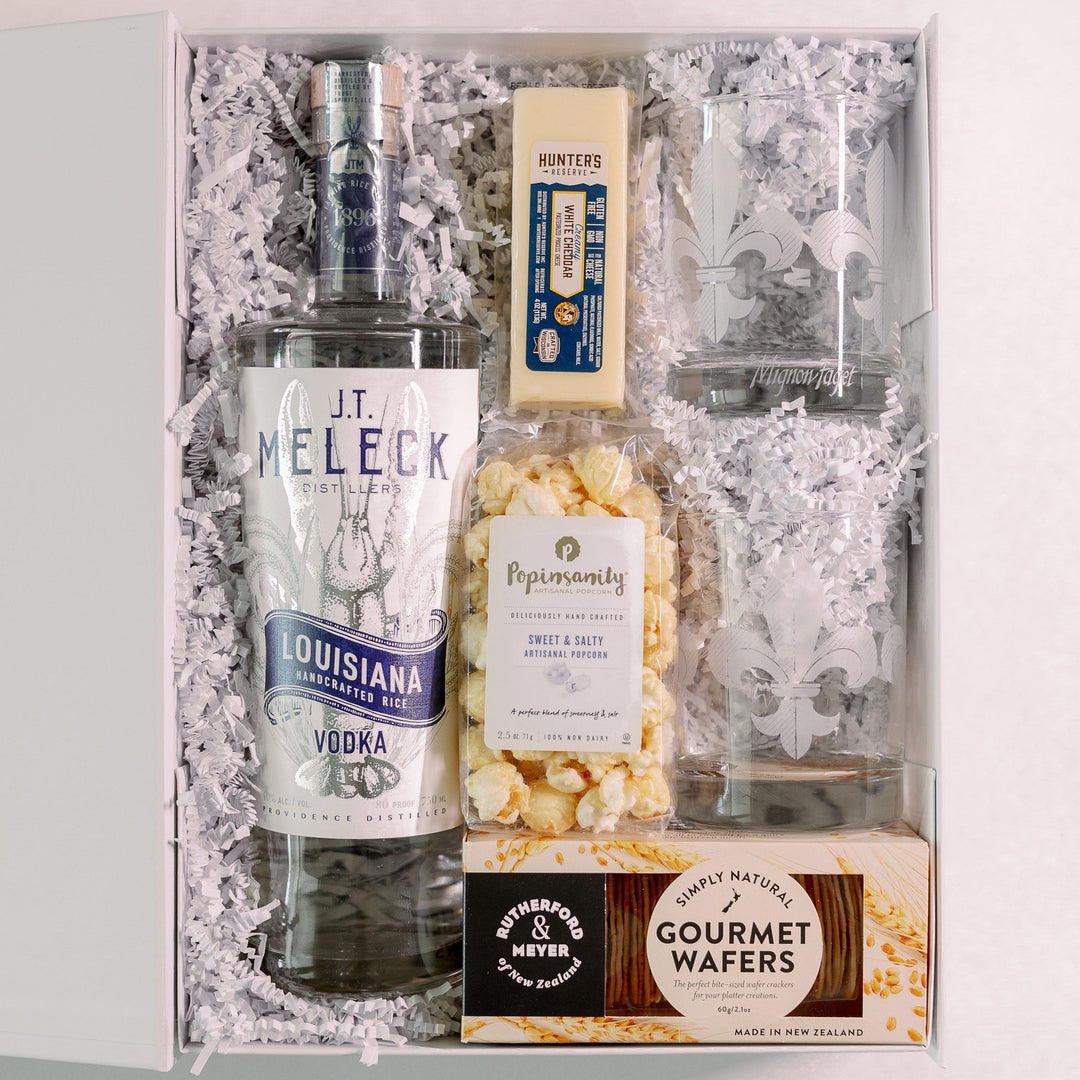 Photograph of a Louisiana-themed gift box featuring J.T. Meleck Rice Vodka and Mignon Faget fleur-de-lis etched glassware presented in white crinkle paper with gourmet white cheddar cheese and sweet and salty popcorn