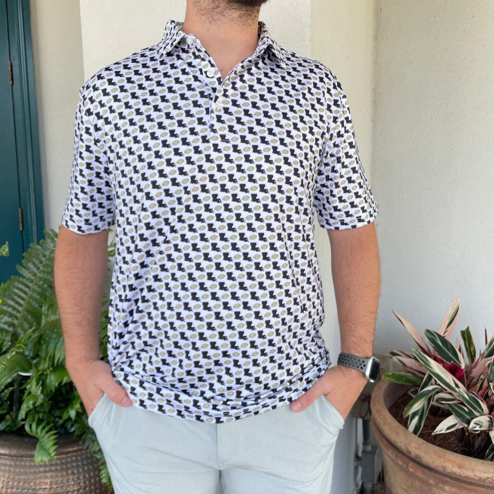 Photograph of a man wearing a white polo shirt featuring a black Louisiana state outline and gold football pattern standing on a porch between a lush green fern and a vibrant pink-striped plant