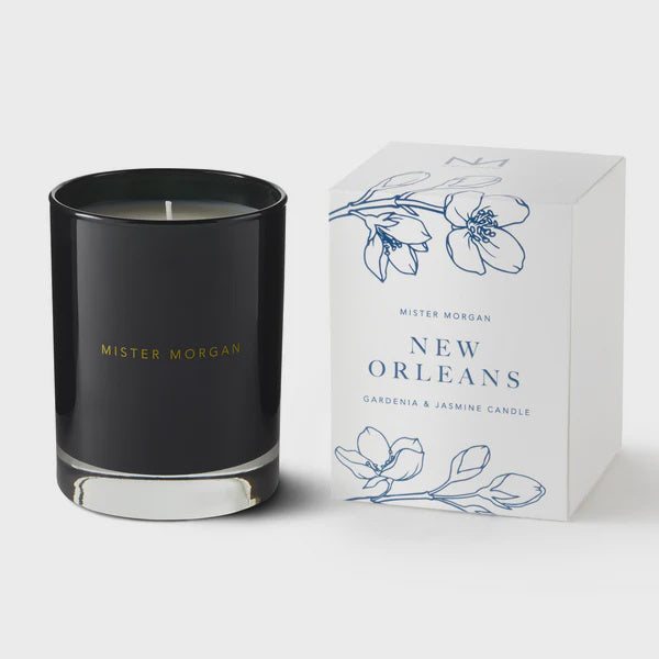 Commercial product photography by Mister Morgan of a New Orleans gardenia and jasmine scented candle in a black glass jar with gold lettering beside its white box decorated with blue floral line art