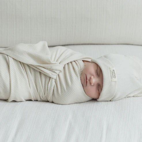 Digital photograph by Good Little Citizen of a serene newborn baby sleeping on a white textured surface swaddled in an off-white ribbed wrap and matching beanie featuring a small rectangular brand label