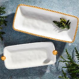 Photograph of porcelain serving trays by Pampa Bay on a blue textured surface featuring two white rectangular platters with a gold beaded rim, spherical gold handles, and fresh green cedar sprigs
