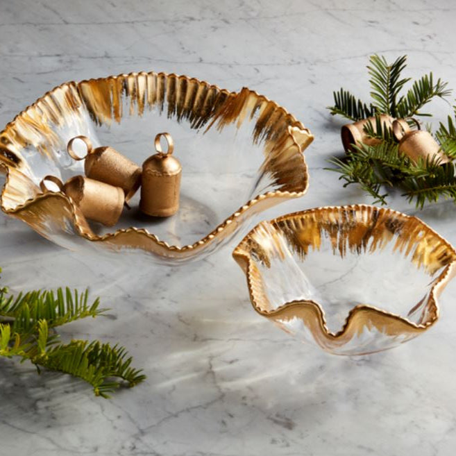 Handcrafted Annieglass clear glass bowls with textured gold-painted rims displayed on a grey-veined marble surface with small brass bells and festive green evergreen sprigs