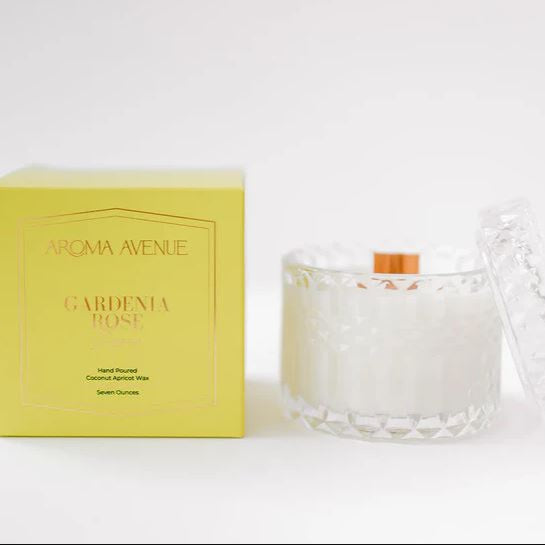 Photograph of an Aroma Avenue Gardenia Rose scented candle in a faceted clear glass jar with a wooden wick beside a bright yellow product box with gold foil lettering on a white background