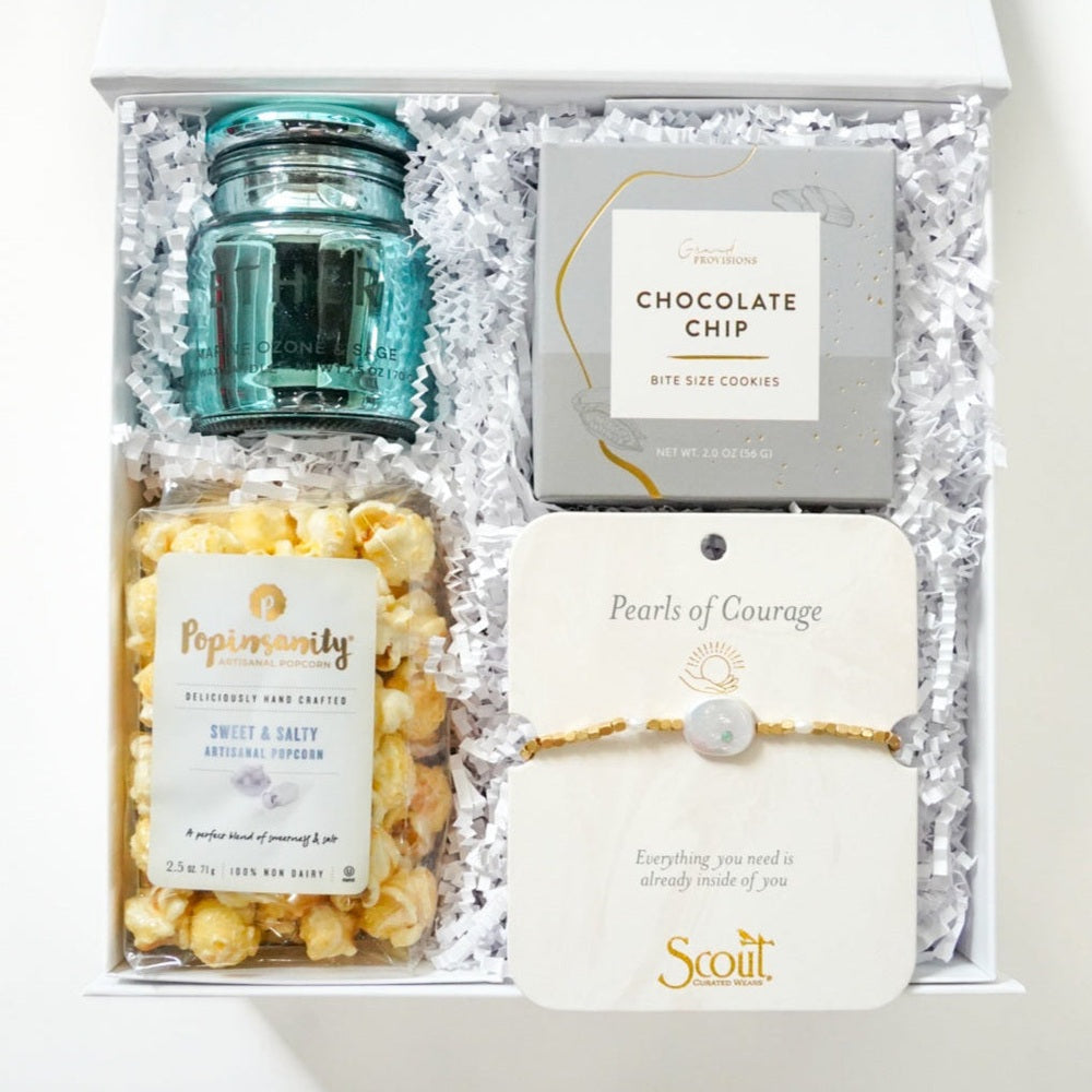 Studio photograph of a curated white gift box containing a teal glass candle, Popinsanity sweet and salty popcorn, Grand Provisions chocolate chip cookies, and a Scout Pearls of Courage gold bead bracelet nestled in white crinkle paper