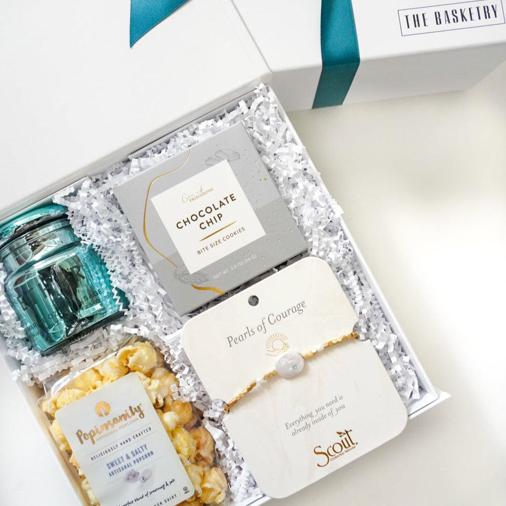 Photograph by The Basketry of a white curated gift box featuring a vibrant teal glass jar, Popinsanity artisanal popcorn, and a Scout Curated Wear gold-toned bracelet nestled in white crinkle paper filler