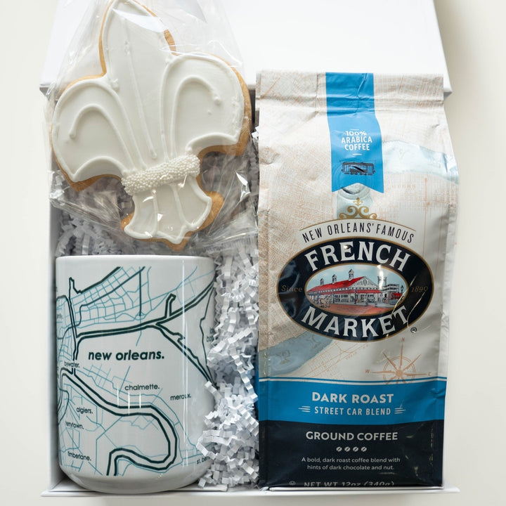 Photograph of a New Orleans themed gift box containing a white fleur-de-lis sugar cookie with beaded icing, a ceramic mug with a teal neighborhood map, and a bag of French Market ground coffee featuring a streetcar logo