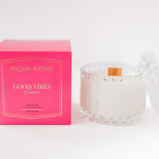 Photograph of Aroma Avenue's Good Vibes Classic candle and packaging on a white surface featuring a bright pink box with gold foil lettering next to a faceted clear glass jar containing white wax and a wooden wick