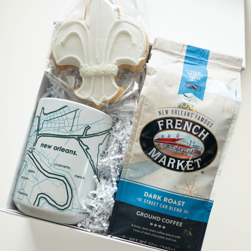 Photograph of a New Orleans themed gift box featuring a white-iced fleur-de-lis cookie with pearl accents, a ceramic mug with a blue map of the Mississippi River, and a bag of French Market dark roast coffee nestled in white shredded paper