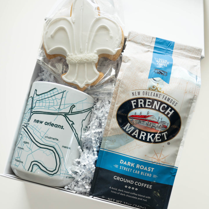 Photograph of a New Orleans themed gift box featuring a white-iced fleur-de-lis cookie with pearl accents, a ceramic mug with a blue map of the Mississippi River, and a bag of French Market dark roast coffee nestled in white shredded paper