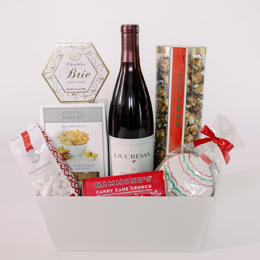 Photograph of a gourmet gift basket on a white background featuring a bottle of La Crema Sonoma Coast wine, a gold-detailed hexagonal Brie cheese box, and a festive frosted holiday cookie tied with a vibrant red ribbon