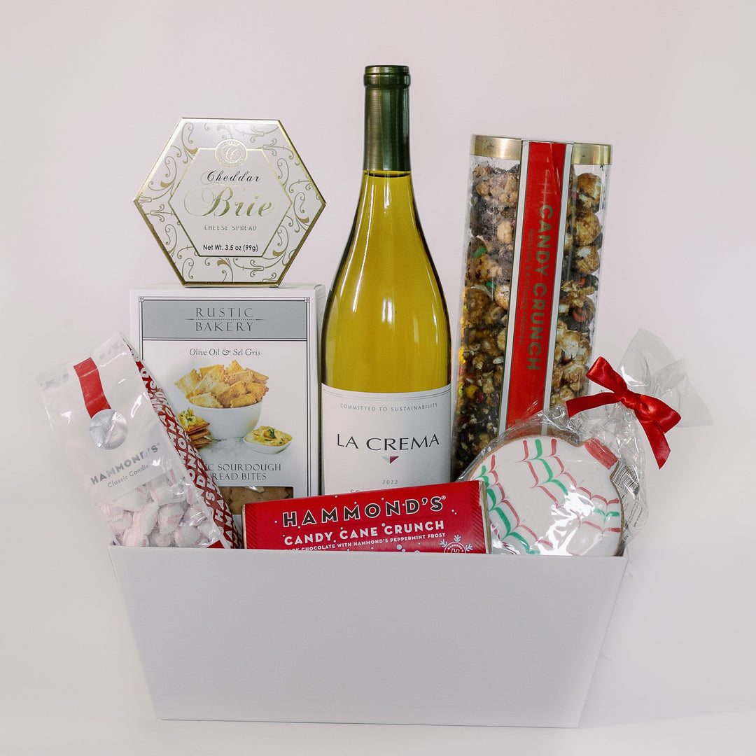 Photograph of a gourmet holiday gift basket in a white studio setting featuring La Crema wine and Hammond's candies including a gold-foiled hexagonal brie box and a festive cookie with red and green icing patterns