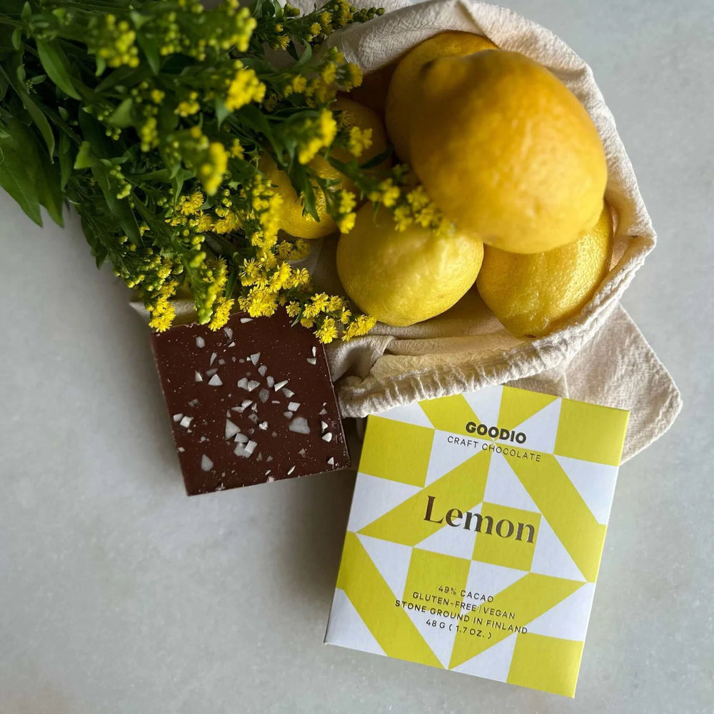 Still life photograph by Goodio featuring a dark chocolate bar with white salt crystals next to its yellow geometric-patterned packaging, fresh whole lemons in a canvas bag, and a bouquet of tiny yellow flowers on a neutral surface