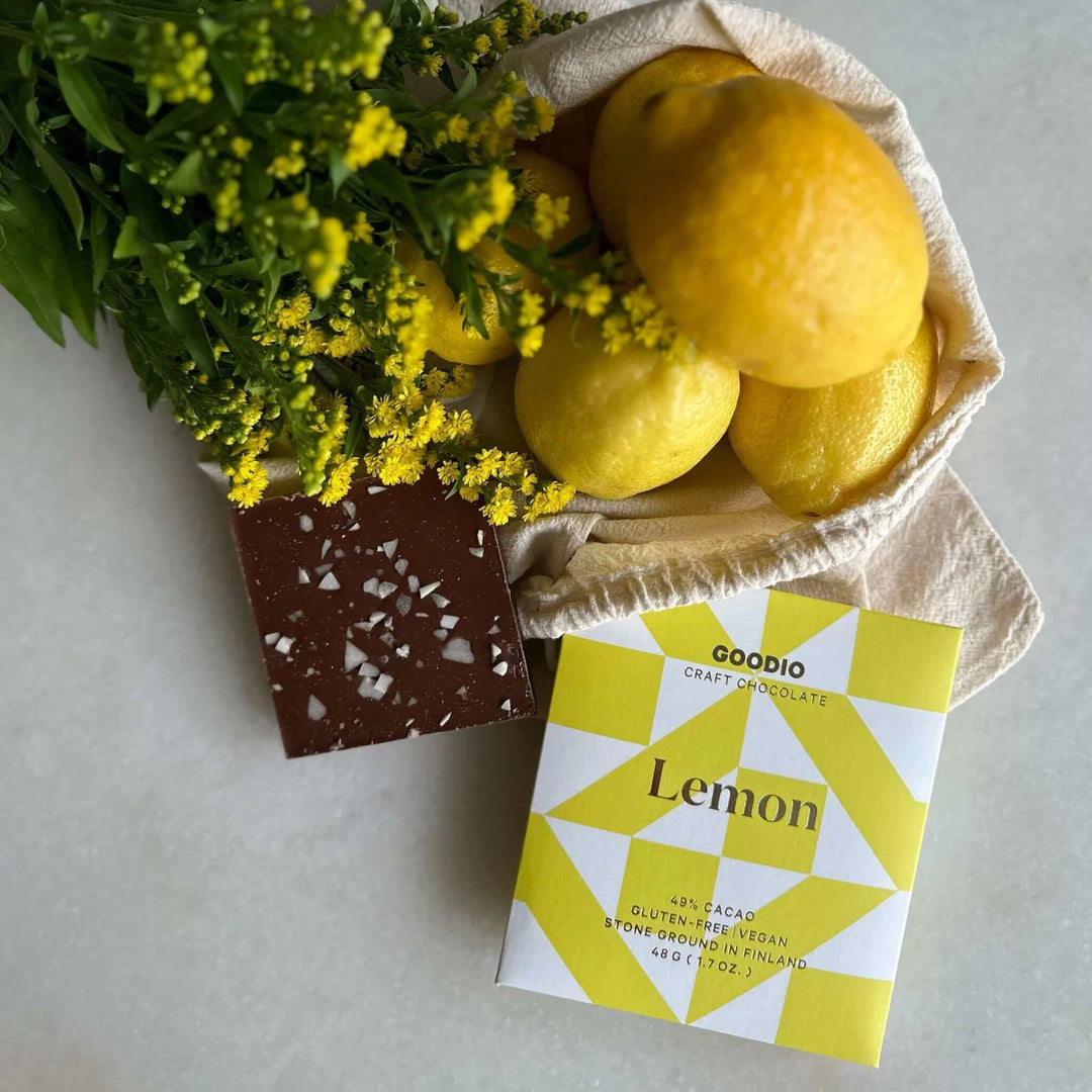 Still life photograph by Goodio featuring a dark chocolate bar with white salt crystals next to its yellow geometric-patterned packaging, fresh whole lemons in a canvas bag, and a bouquet of tiny yellow flowers on a neutral surface