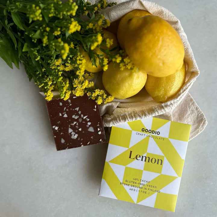 Still life photograph by Goodio featuring a dark chocolate bar with white salt crystals next to its yellow geometric-patterned packaging, fresh whole lemons in a canvas bag, and a bouquet of tiny yellow flowers on a neutral surface