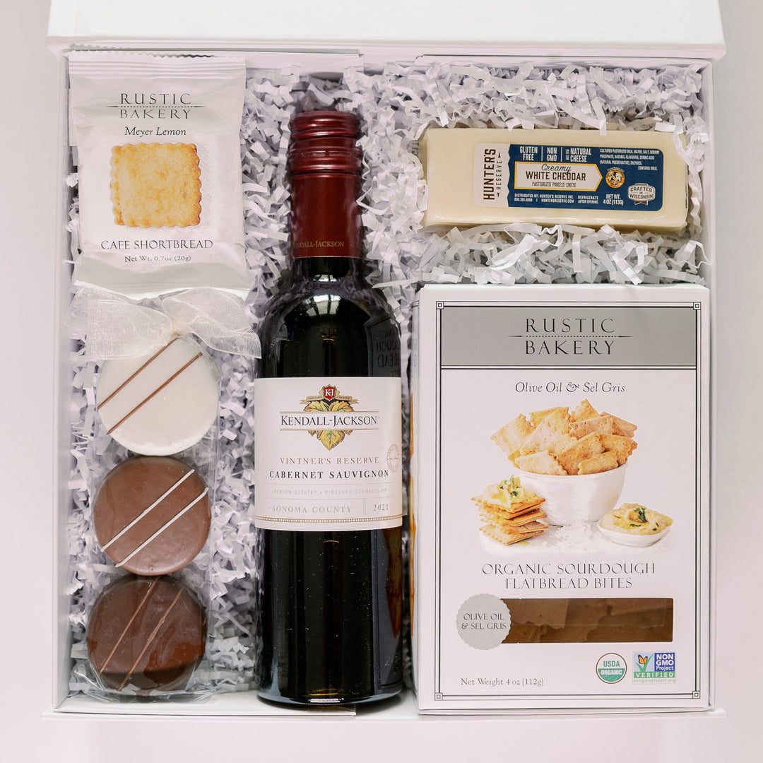 Photograph of a Kendall-Jackson gourmet gift box from Sonoma County featuring a 2021 Cabernet Sauvignon with a maroon foil cap, Hunter’s Reserve white cheddar, and Rustic Bakery crackers nestled in white shredded paper with three drizzled chocolate cookies tied in a white bow