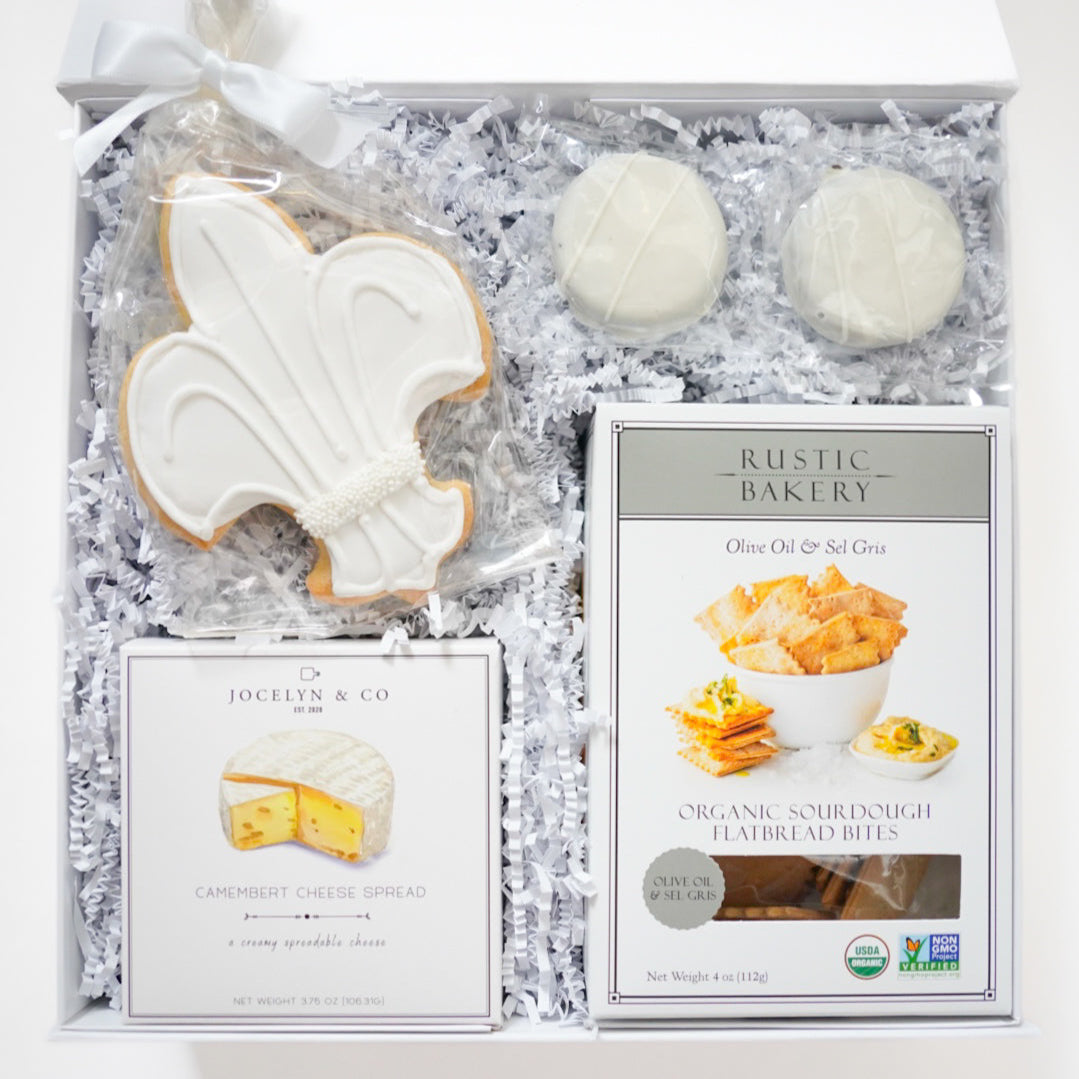 Photograph of a curated gourmet gift box by Jocelyn & Co and Rustic Bakery featuring a large white fleur-de-lis iced cookie, organic sourdough flatbread bites, and Camembert cheese spread nestled in white crinkle paper with a satin ribbon bow