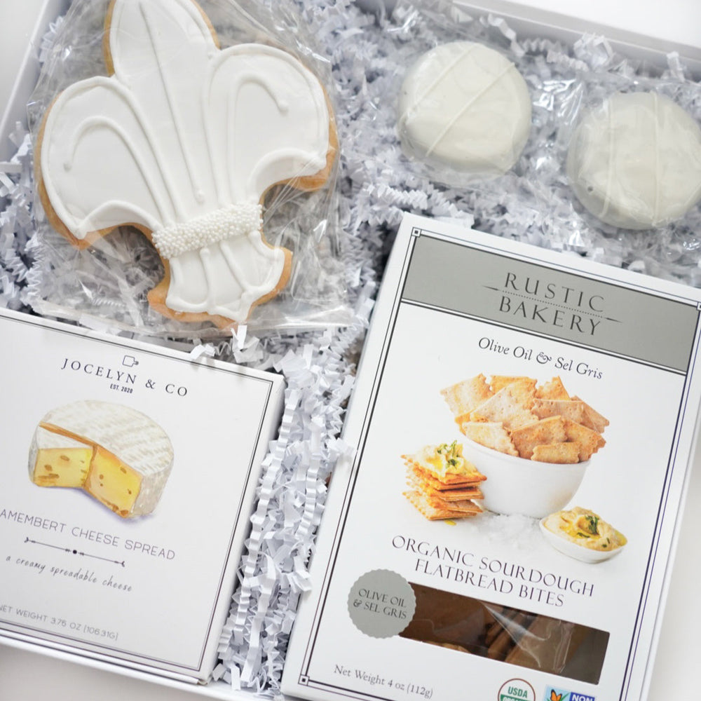 Product photography of a gourmet food gift box featuring a large white fleur-de-lis sugar cookie with decorative pearl sprinkles, Jocelyn & Co Camembert cheese spread, and Rustic Bakery sourdough flatbread bites nestled in white crinkle paper shred