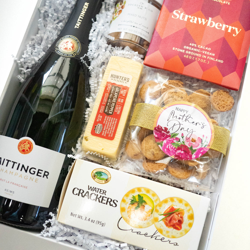 Photograph of a Mother's Day gift box featuring a bottle of Taittinger Champagne, a block of jalapeño pepper cheese, a vibrant red strawberry chocolate bar, and cookies decorated with a gold ribbon and a pink floral sticker
