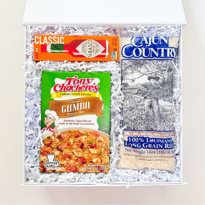 Photograph of a Louisiana-themed gift box featuring a bright green Tony Chachere’s Gumbo mix, a signature red Tabasco sauce bottle, and a bag of Cajun Country rice showcasing a traditional accordion player illustration within white crinkled paper filler