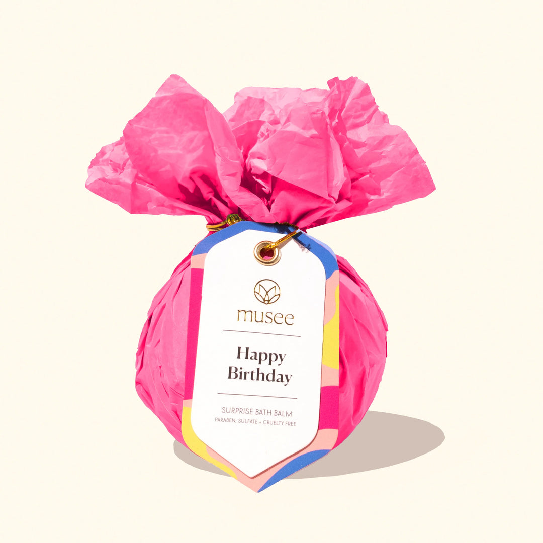 Product photograph by Musee of a surprise bath balm wrapped in ruffled hot pink tissue paper featuring a colorful Happy Birthday tag and gold string on a cream background