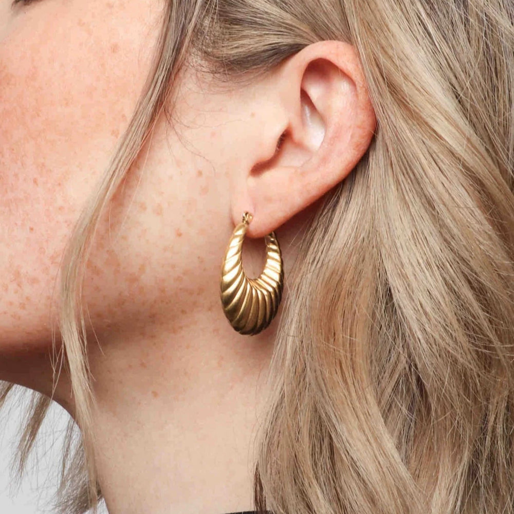 Photograph of a woman's ear featuring a textured gold croissant-style hoop earring, framed by loose blonde hair and delicate sun-kissed freckles