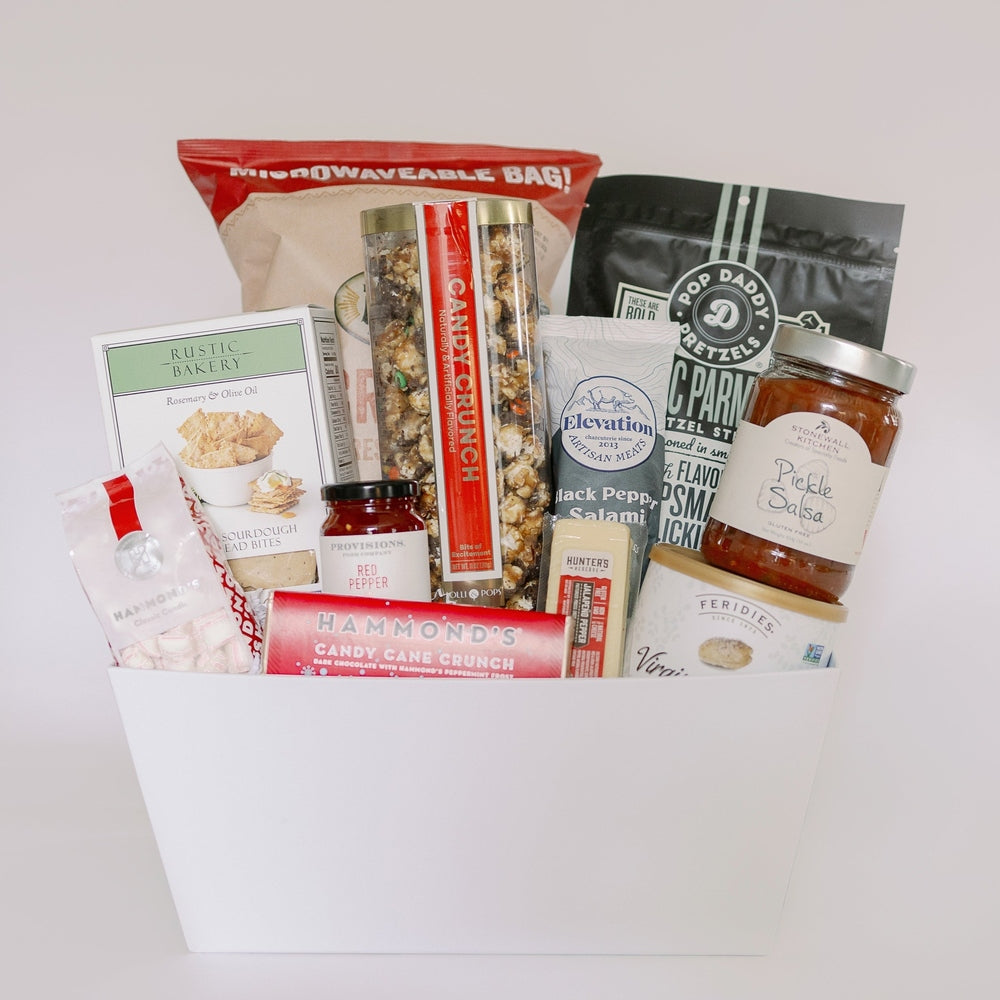 Photograph of a white gourmet snack gift basket featuring a tall tube of Hammond's Candy Cane Crunch, a jar of Stonewall Kitchen Pickle Salsa, and Rustic Bakery sourdough flatbread bites against a plain studio background