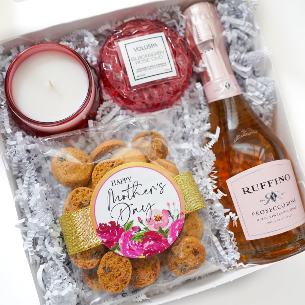 Photograph of a curated Mother's Day gift box featuring a bottle of Ruffino Prosecco Rosé and a red faceted Voluspa candle alongside a bag of chocolate chip cookies decorated with a gold glittery ribbon and a pink floral sticker nestled in white shredded paper