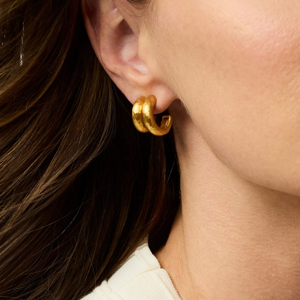 Photograph of thick double-banded gold hoop earrings with a textured hammered finish and warm yellow glow worn on a woman's earlobe near her neckline and dark wavy hair