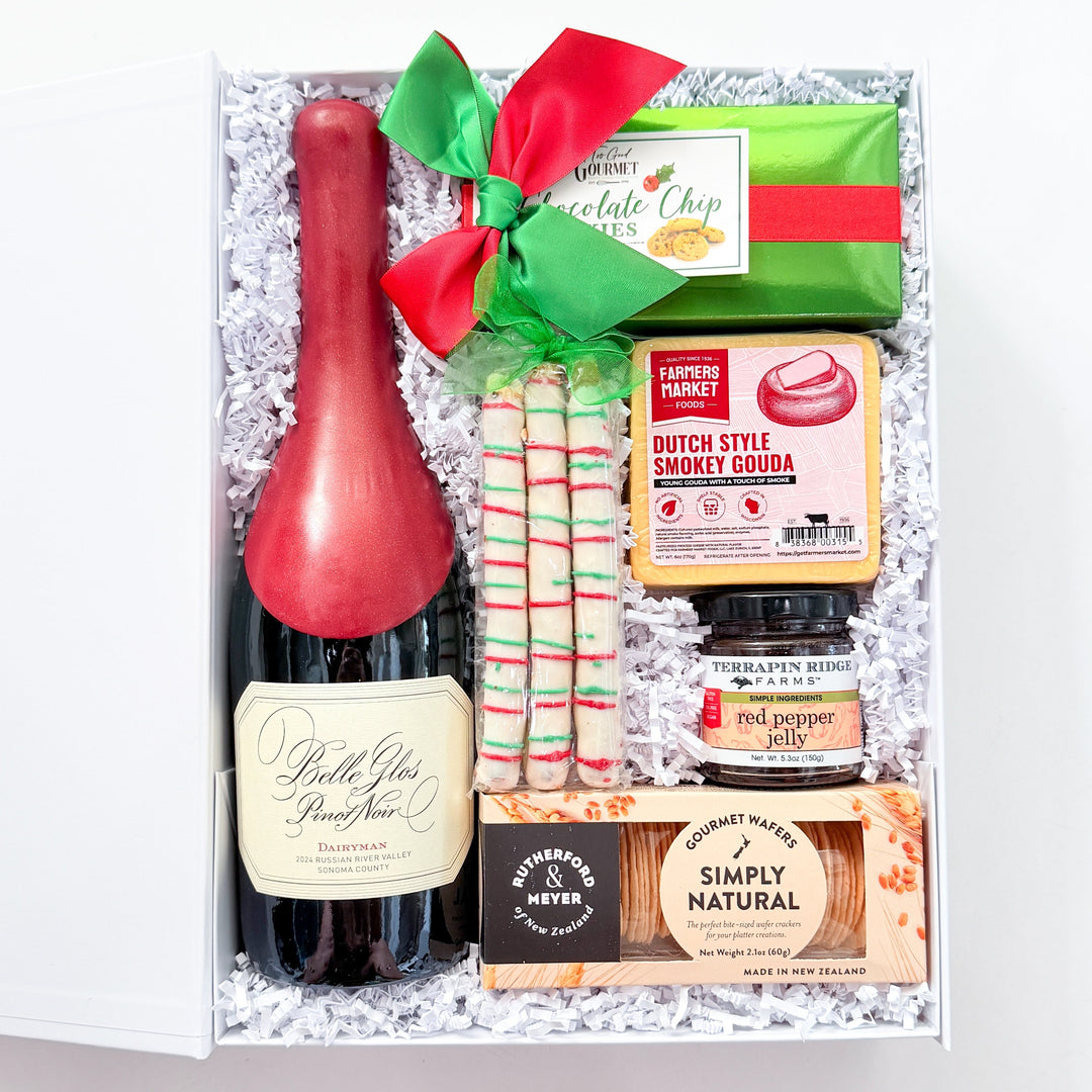 A curated gift box containing a bottle of Bell's Vie Fraiche wine, red and white striped candy canes, a green chocolate chip cookie, a jar of Dutch Style Candy Cane Soup, and a bar of Simply Natural chocolate, all arranged in white crinkle paper with a red and green bow.