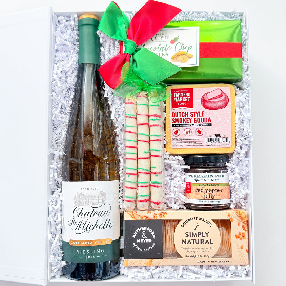 A curated gift box set containing a bottle of Chateau de Michelle Riesling, Dutch Style Sweet Cobra candy canes, a chocolate chip cookie, a jar of pepperoni, and Simply Natural crackers, arranged in white crinkle paper with a green and red ribbon.