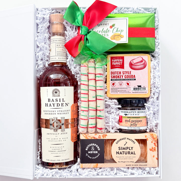 A curated gift box set on a white background, featuring a bottle of Basil Hayden bourbon, red and white striped candy canes, a green chocolate chip cookie, a box of Dutch Style Pancake Flour, a jar of red pepper flakes, and a package of Simply Natural crackers, all arranged in white crinkle paper with a red and green bow.
