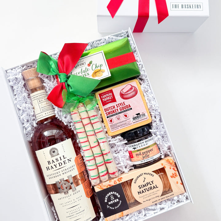 A curated gift box set on a white background, featuring a bottle of Baileys Irish Cream, a green and red striped striped sweater, a bag of Simply Natural chocolate, and other gourmet snacks, arranged in white crinkle paper next to a white gift box with a red ribbon.