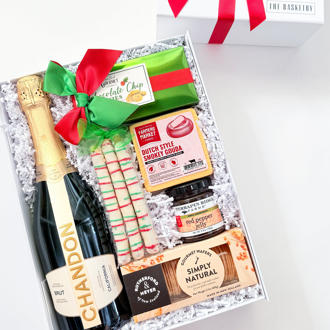 A curated gift box set on a white background, featuring a bottle of Chandon sparkling wine, a green and red striped candy cane, a bag of Simply Natural graham crackers, a jar of chocolate spread, and a white gift box with a red ribbon.
