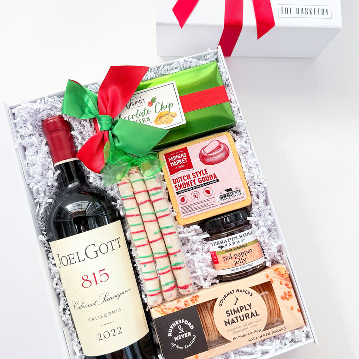 A curated gift box set from The Blackstone featuring a bottle of red wine, candy canes, gourmet snacks, and a white gift box with a red ribbon, arranged in white crinkle paper.